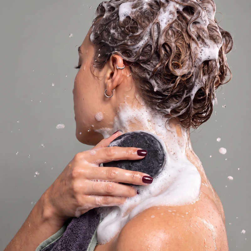 A woman with curly brown hair covered in shampoo foam is shown from the side and back, using a Protect & Repair Shampoo Bar to lather her neck and shoulder against a plain gray background.