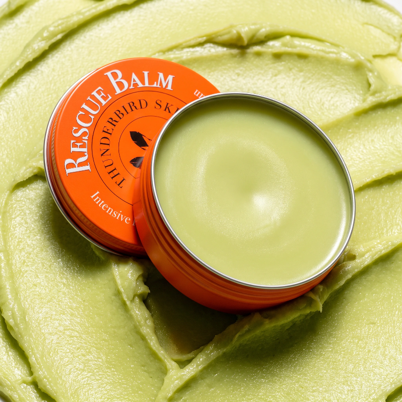 An open round tin of Rescue Balm with a bright orange lid resting beside it on a textured light green balm surface. 