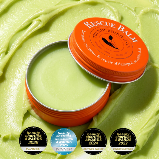 An open round tin of Rescue Balm, showing the light green balm inside, with its lid leaning against it. The tin has a bright orange lid with white text and shows the awards it has won.