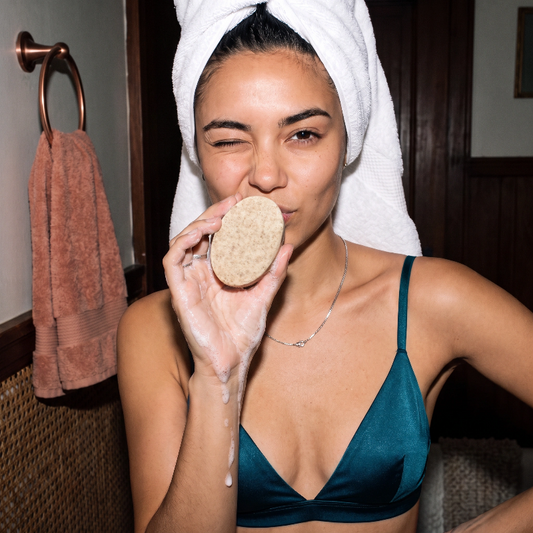A woman with a white towel wrapped around her head holds a Skin SOS Cleansing Bar near her face, wearing a teal satin bra in a bathroom setting.