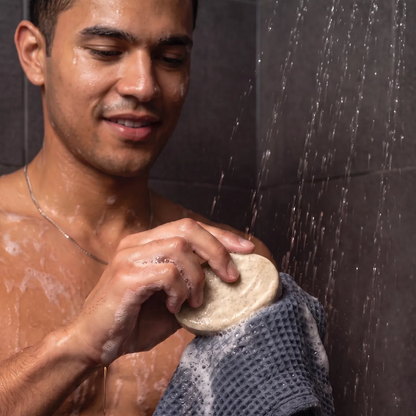 A shirtless man standing in a shower under running water, holding a Skin SOS Cleansing Bar.