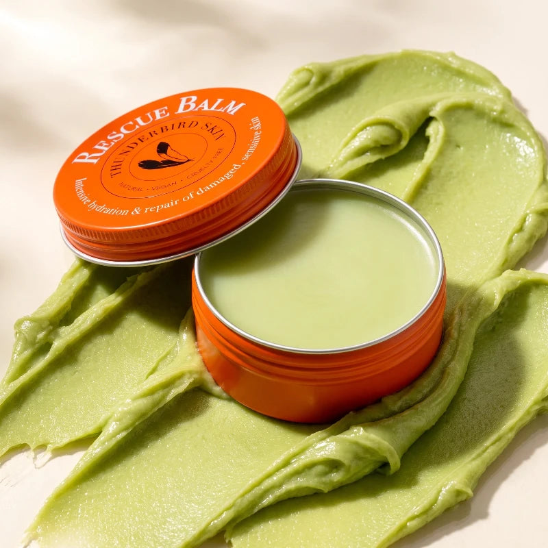 An open round tin of Rescue Balm with its orange lid resting beside it, set on top of a thick smear of pale green balm. The tin has a bright orange lid with white text reading 'RESCUE BALM'.
