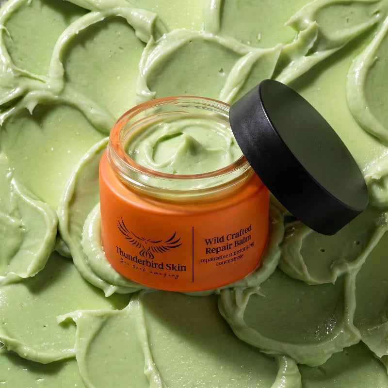 An open jar of Thunderbird Skin 'Wild Crafted Repair Balm' with its black lid resting beside it, set against a background of swirled light green balm cream.